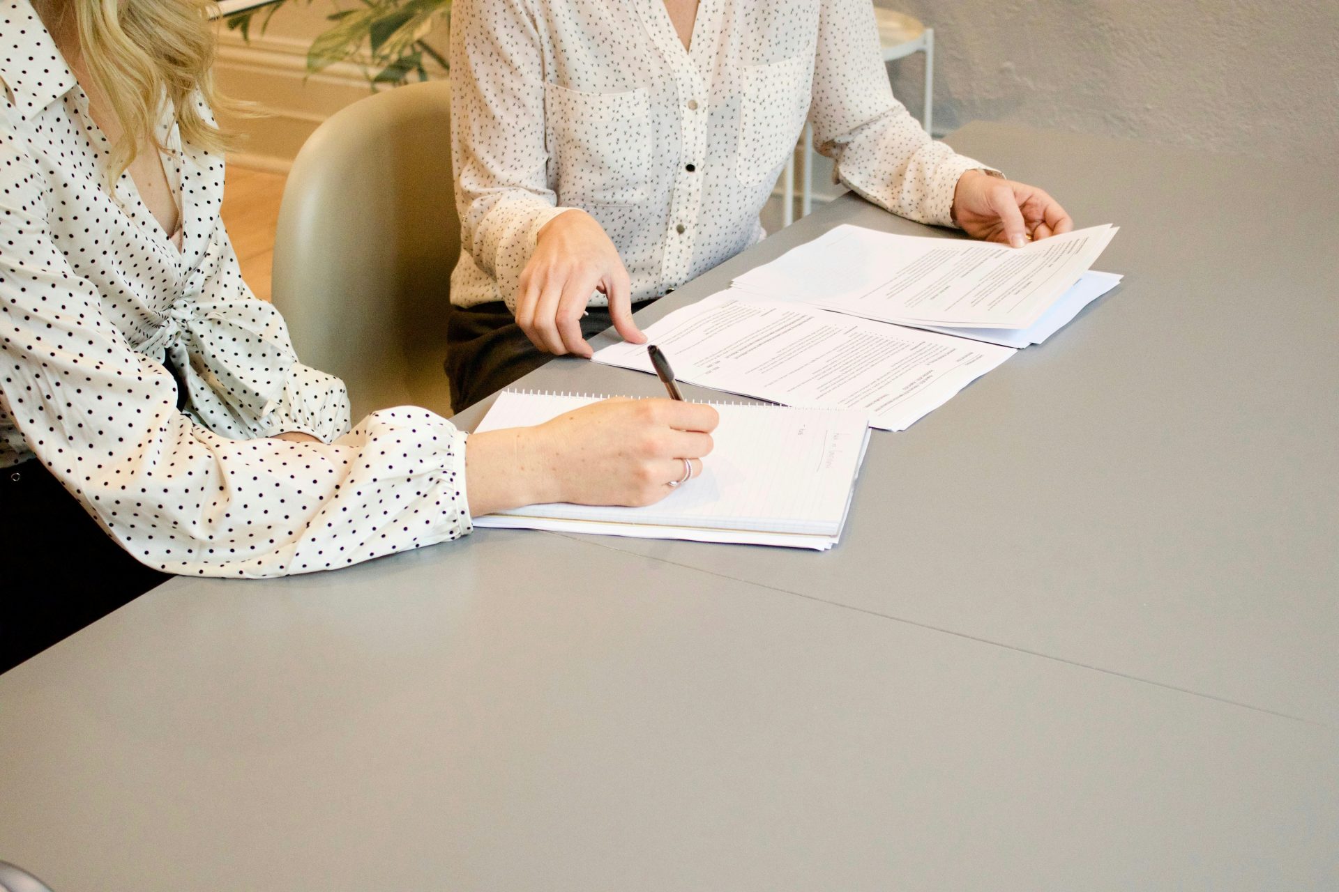 Une femme signant sur une feuille de papier blanc, à côté d'une autre femme sur le point de toucher les documents.